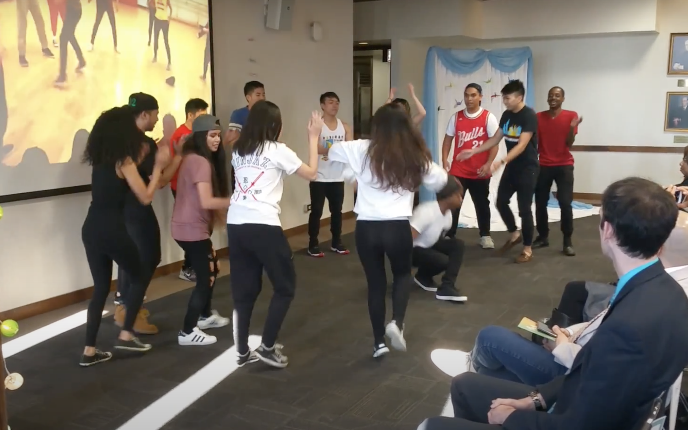 One circle of dancers of many races surrounds one mixed Black and Asian man dancing in the center